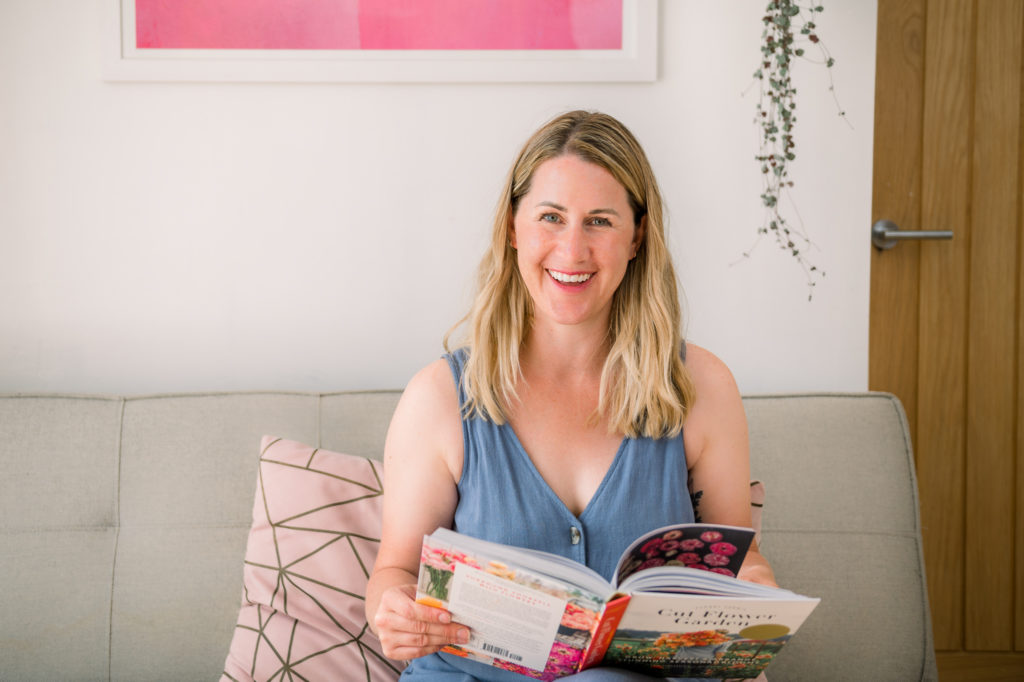 Blonde haired female business owner sitting on a sofa looking to camera whilst holding a book 