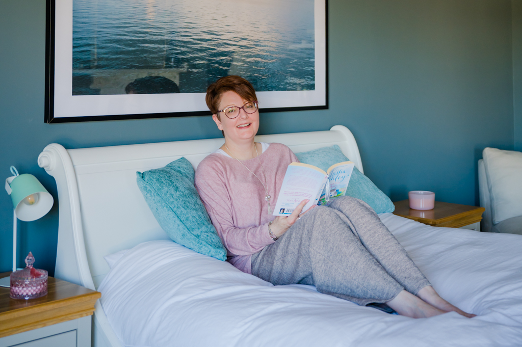 Woman with auburn hair lying on a bed with a book smiling