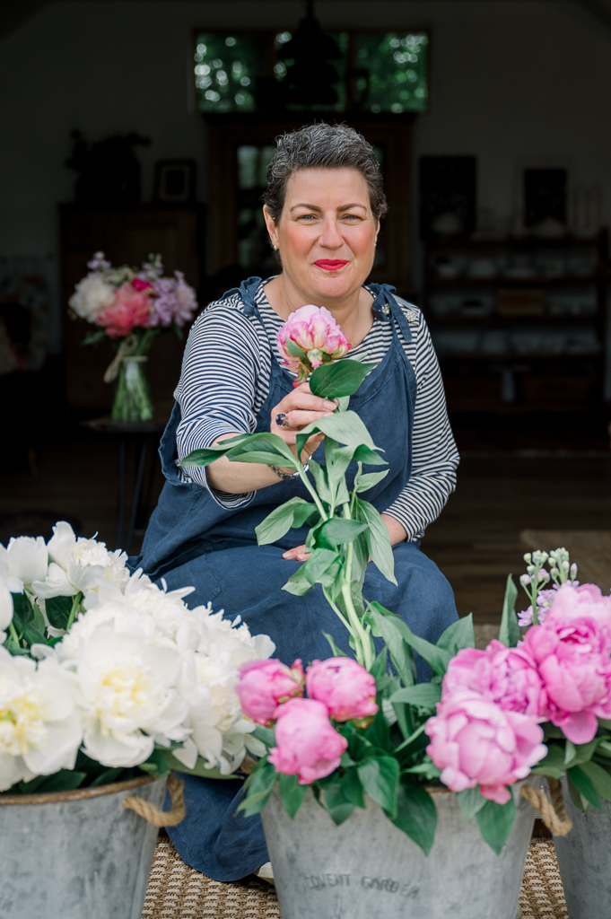 Woman with short hair sitting in front of flowers holding a pink peony