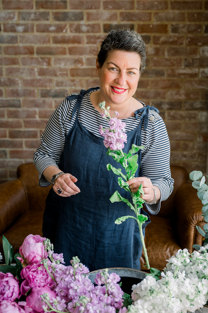 Smiling woman wearing a stripey top and dungaree dress holding a flower with more flowers surrounding her