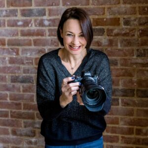 Smiling woman in a black sparkly jumper holding a camera and smiling with a brick wall in the background