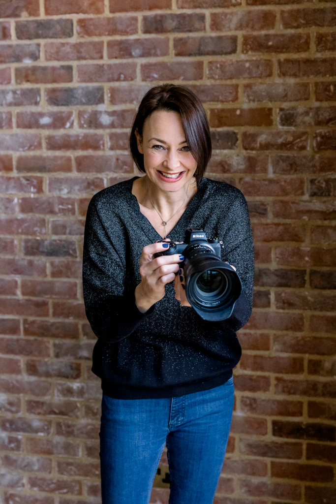 Brown haired smiling woman standing up holding a camera