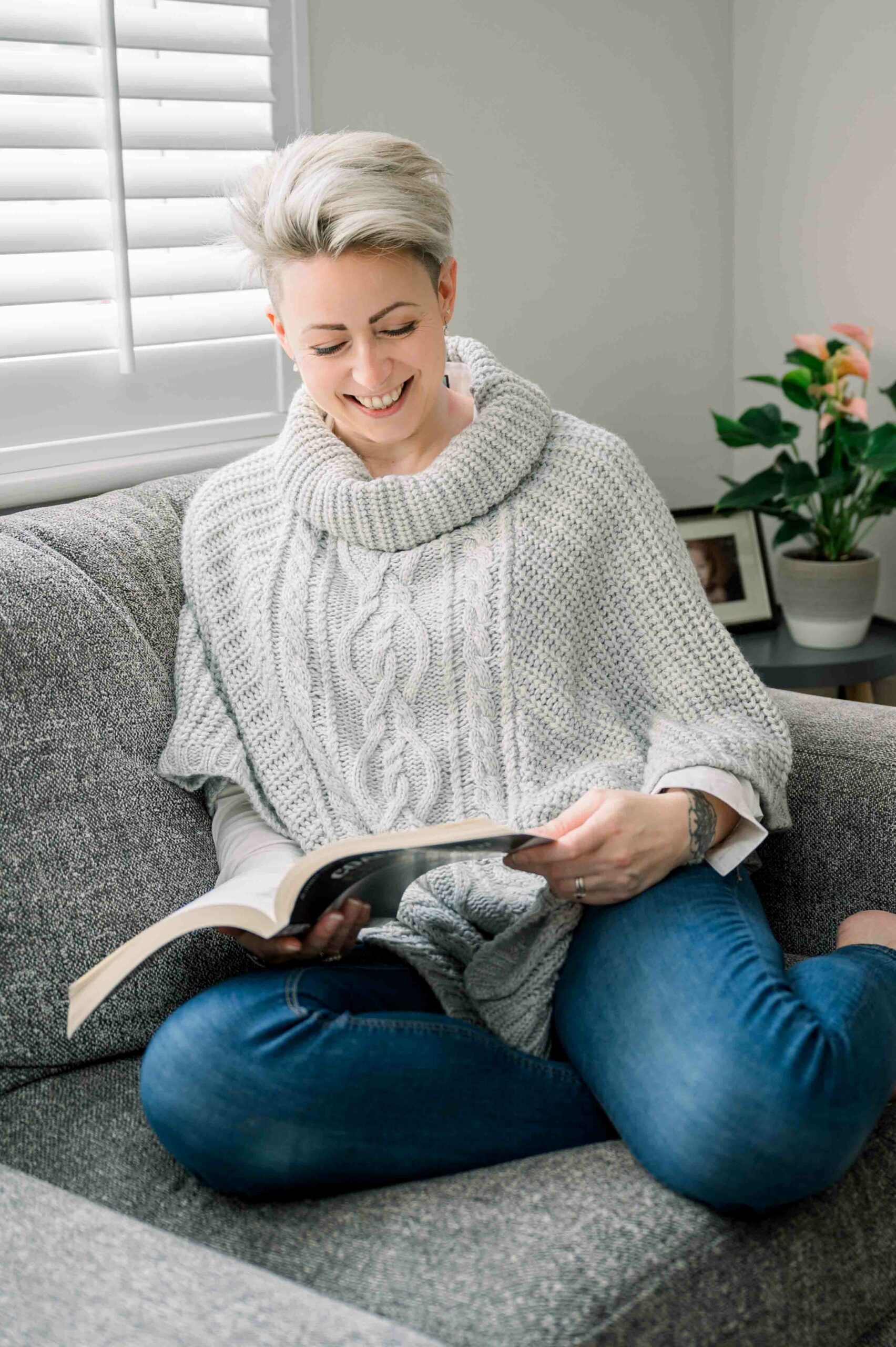 smiling woman with short blonde hair sitting on her sofa reading a book