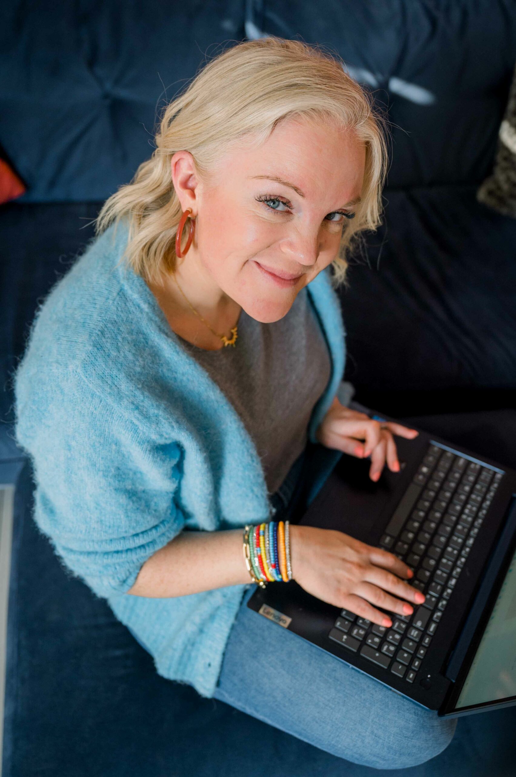 Blonde haired woman working with a laptop on her lap looking up to camera