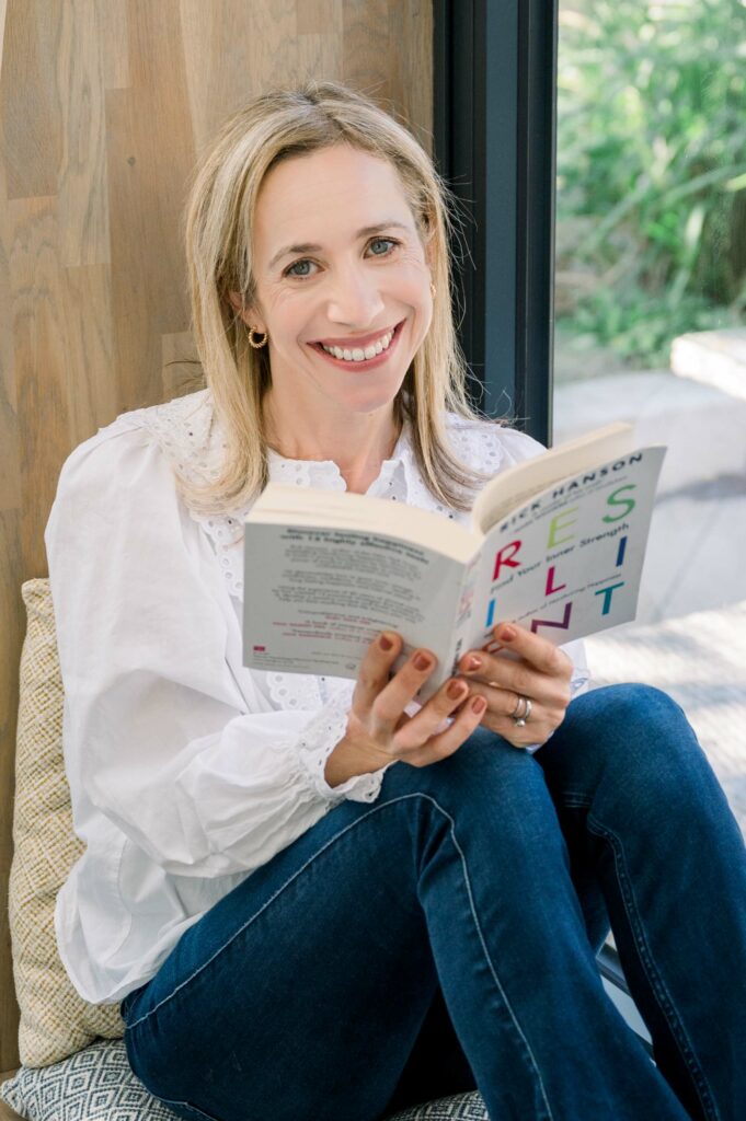 Smiling lady with blond hair sitting with her knees up, holding a book and looking at the camera