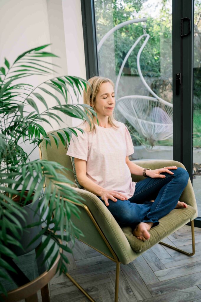 Lady sitting in a green armchair with close eyes and crossed legs with a large plant next to her